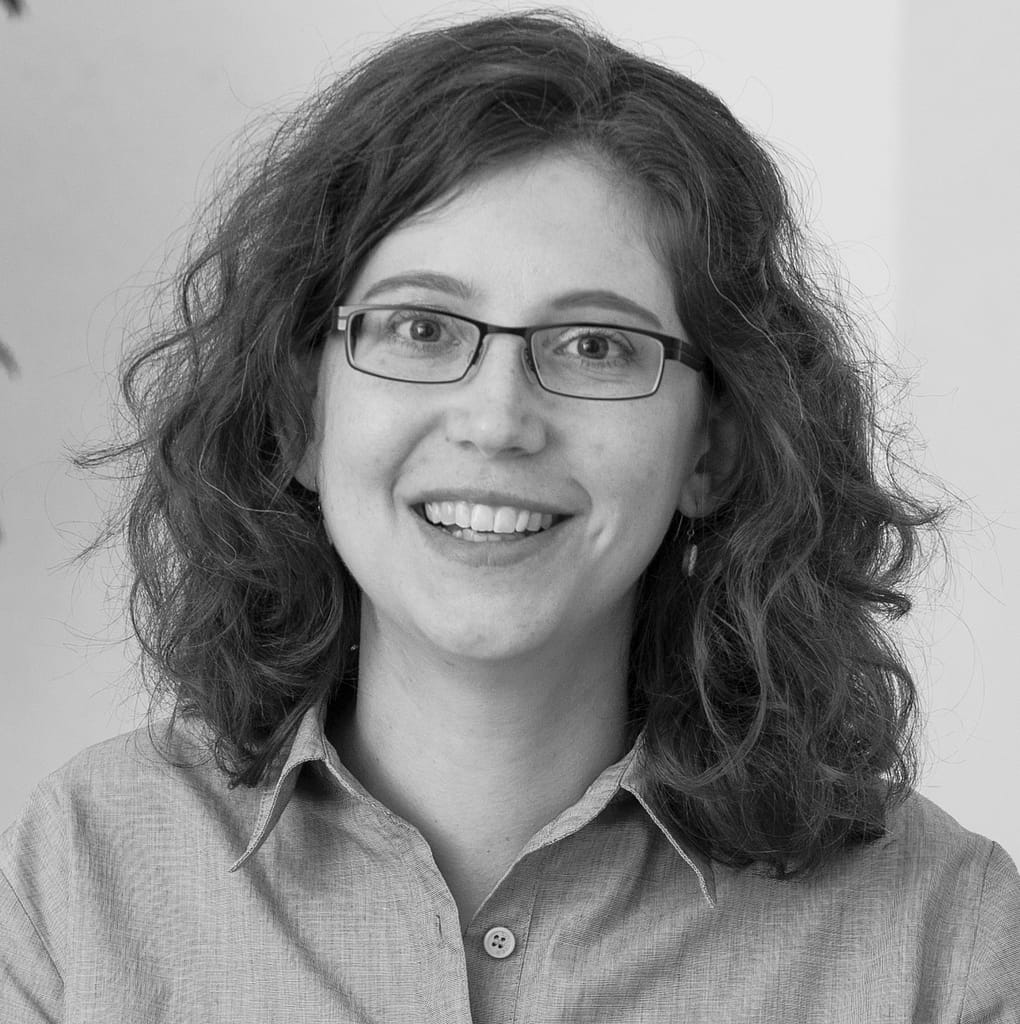 Black-and-white portrait of a smiling white person with curly brown hair and glasses.