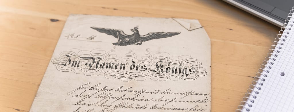 Ein Urteilsspruch aus der Kaiserzeit liegt auf einem Schreibtisch; der Titel lautet "Im Namen des Königs".