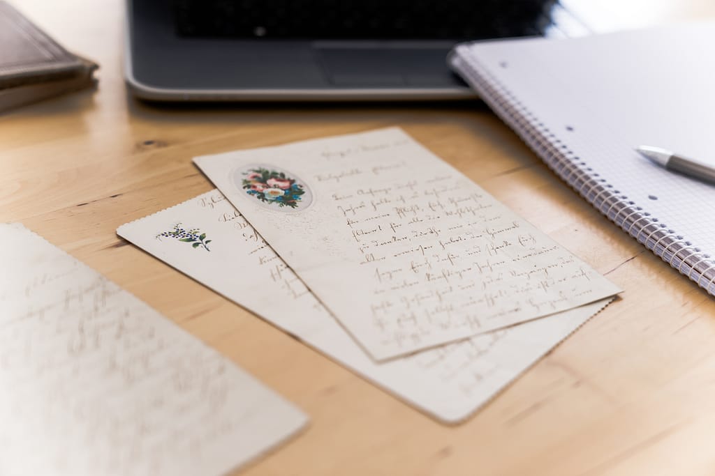 Two old letters are lying on a wooden desk. At the side and in the background there are a noteblock and a laptop.