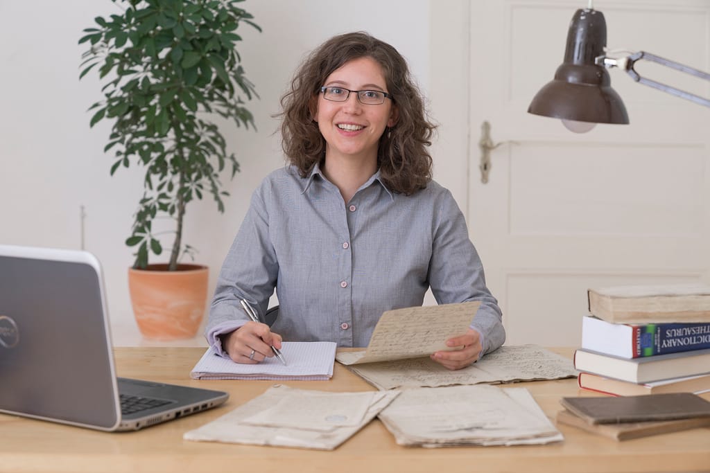 Eine weiße Person mit Brille und lockigen braunen Haaren sitzt an einem Schreibtisch; sie hat einen Laptop, Bücher und mehrere alte Dokumente vor sich liegen. Sie schaut in die Kamera und lächelt.