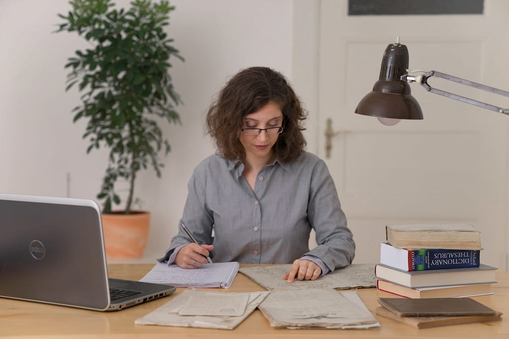 A white person with brown curls and glasses is sitting at a desk, focused on her work. In front of her are a noteblock and some old documents, as well as a laptop and several books.