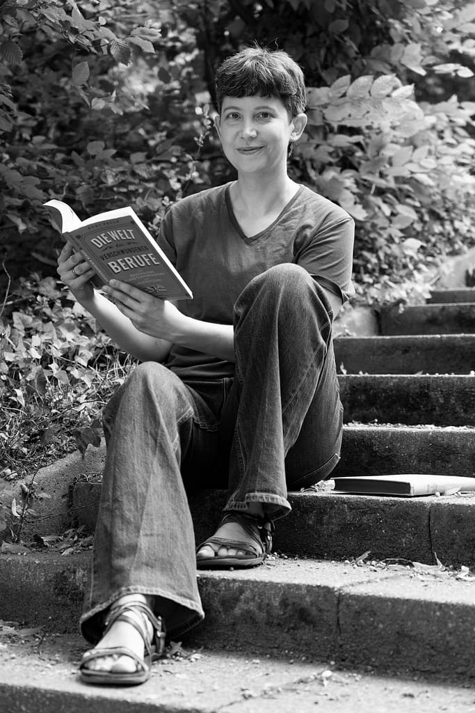Arin Dachner sitzt auf einer Treppe in einem Park und sieht lächelnd von einem Buch über alte Berufe auf.